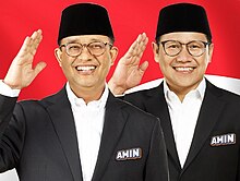 Picture of two man, Anies Baswedan on the left and Muhaimin Iskandar on the right wearing black cap and suit saluting in front of waving Indonesian flag.