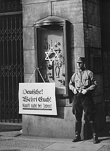 Photographie en noir et blanc d'un membre de la SA à côté d'une affiche proclamant : « Allemands ! Défendez-vous ! N'achetez pas chez les Juifs ! » en 1933