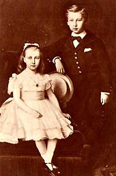 Henriette, portant une robe claire évasée, les cheveux retenus par un bandeau est assise à côté de son frère debout en costume sombre avec une cravate et légèrement appuyé sur le fauteuil où se tient sa sœur