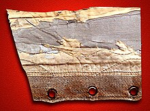 A piece of badly distressed fabric on a red background. The fabric is crossed by three seams and there are three eyelets at the bottom of it.