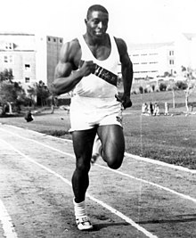 Foto em preto e branco de um atleta, de frente, correndo em uma pista cinzenta.