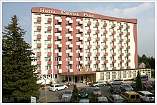 Zdjęcie przedstawia budynek hotelu Cristal Park. Budynek jest wysoki, pomalowany na biało i czerwono. W bryle budowli znajduje się wiele okien i małych balkonów. Po lewej stronie budynku rośnie wysokie iglaste drzewo, a na ziemi widoczny jest parking pełen samochodów.