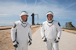 Os astronautas da NASA Douglas Hurley (à esquerda) e Robert Behnken (à direita) com o Falcon 9 atrás deles com o ônibus Crew Dragon instalado no topo, na Plataforma de Lançamento 39A