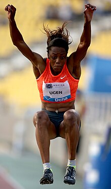 Black women in orange top jumping with arms in air.