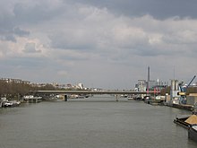 Fotografie a podului din aval care traversează Sena.  În dreapta o barjă care transportă materiale și o zonă industrială