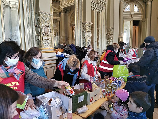 People in an ornate building handing out cartons and cans of food with a pile of clothes in the background