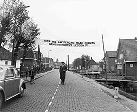 Burgemeester van Landsmeer in 1958 onder spandoek met tekst: 'Hier wil Amsterdam haar nieuwe gemeentegrens leggen'. Sinds 1966 is hier een paar meter vandaan de huidige gemeentegrens van Kadoelen (Gemeente Amsterdam) en Gemeente Landsmeer.