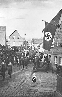 Photographie en noir et blanc montrant le défilé de charrettes ornées dans la petite rue d'un village pavoisé de croix gammées.