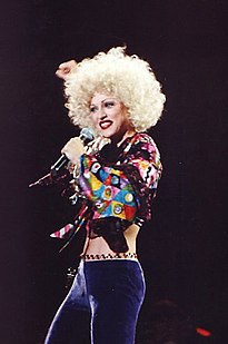 Image of a blond female facing her left. She's wearing purple pants and a maroon blouse with different color squares on the sleeve. She's also wearing an afro hairstyle and with her right arm she holds a microphone to her mouth and has her left arm lifted back above her head.