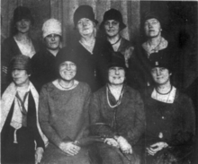 A group of nine white woman, most smiling and wearing hats; four seated in the front row, and five standing in the back row.