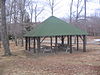 A green pyramidal roof supported by log pillars over three picnic tables, leafless trees are around it and a road runs behind