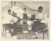 Black and white photo of two Black men and a white woman under a large tent. All three are sitting at a wood desk covered with stacks of books; the woman is typing on a typewriter.