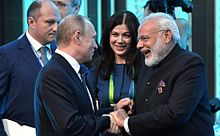 Two standing men are pictured shaking hands. The first is dressed in Indian clothing; the second is in a Western business suit; both standing behind a Russian flag.