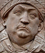 bas relief : portait de Trithemius, un homme coiffé d'une toque ornée de tresses et de feuilles, fossette au menton, relève les sourcils et plisse le front.