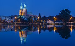Katedra gnieźnieńska nocą i jej lustrzane odbicie w wodach jeziora Jelonek. W tle budynki starego miasta.