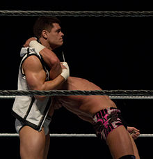 Two Caucasian males in a wrestling ring. A black-haired male wearing white wrestling tights and a black and white jacket is standing, with an inverted headlock on the other who is bent over backwards.