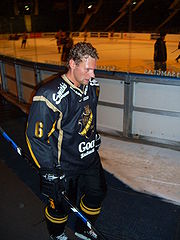 An ice hockey walking to the right of the camera. Behind is the ice hockey rink. He has short brown hair and is not wearing a helmet. He is wearing a black uniform.
