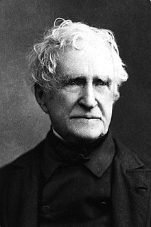 Black-and-white portrait photograph of an old white man with wavy white hair and wearing a black coat