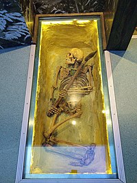Skeleton in a glass casket viewed from above, clutching a spear