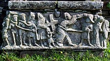 The procession depicting Jesus carrying the cross. On the left we see the good and bad robbers roped together around the neck, the end of the rope being held by a soldier who wears a chapeau à bec, a form of headgear popular in the reign of Louis XI of France. The next carving depicts another soldier who leans on the cross to make it heavier but Simon of Cyrene, shown as a very small figure, is under the cross and tries to lift it to counter the soldier's action. Jesus Christ comes next, also with a rope around his neck and led by another soldier. He carries a hammer which will be used to nail Christ to the cross. Another soldier comes next and finally we see John the Evangelist trying to comfort the Virgin Mary as they both lead the procession towards Golgotha