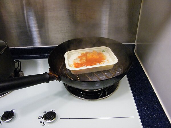 A makeshift steaming vessel with lid removed; a frozen dish is placed on a metal frame in a single handled wok with water.