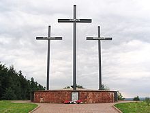 La photographie couleur représente une allée dallée qui conduit à une surélévation maçonnée supportant trois croix métalliques. Chacune porte le nom des massacres subis par les Polonais : Katyń, Charkow et Miednoje. Devant le mémorial, un bas relief est fleuri par trois gerbes de fleurs rouges et noires.