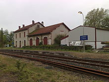 Photographie de deux bâtiments aux boiseries rouges sang, en bordure d’une voie ferrée. Un panneau sur fond bleu indique Ustaritz/Ustaritze.
