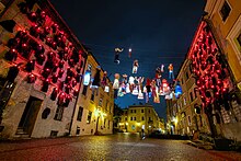 Fotografia przedstawia instalację artystyczną zawieszoną w przestrzeni Starego Miasta w Lublinie. Zdjęcie wykonane jest nocą. Ulica Grodzka, dwie przeciwległe kamienice, pomiędzy nimi rozwieszone liny, na których zawieszone są kolorowe podświetlone stroje. Na ścianach kamienic wiszą natomiast czarne futra, z kołnierzy wystają dzioby-maski przypominające maski lekarzy podczas epidemii dżumy. Maski mają czerwone świecące oczy.