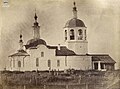 Photographie en noir et blanc de face de l'église de l'Exaltation de la Sainte Croix au cimetière Sevastianovskoïe. L'église possède un clocher principal à droite et un clocher secondaire à gauche. Au premier plan, on distingue des croix des tombes d'un cimetière, avec une petite clôture faite de poteaux de bois et de fils.