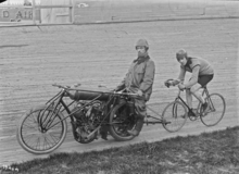Cycliste en piste sur un vélodrome, derrière une moto.