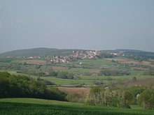 Un pequeño pueblo con una torre de iglesia visible, en una colina baja rodeada de campos y árboles, visto desde otra colina similar