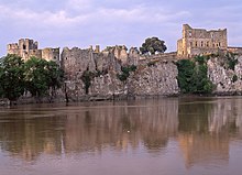 .Château de vielles pierres en ruine, formé de murs bruts et de larges tours, s'étalant en longueur au sommet d'une petite falaise, au pied de laquelle coule une rivière. Le ciel est bleu pastel et le château est rosé, dû à l'heure matinale à laquelle la photo a été prise.