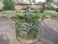 Photograph of a formal garden at the Luther Burbank House and Gardens, featuring brick walkways and planterboxes, a small lawn, a fountain and pool, low stone walls, and wooden trellises.