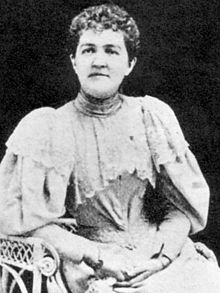 Femme assise dans un fauteuil, cheveux châtain coupés court et frisés.