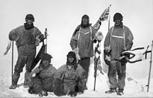 Five men(three standing, two sitting on the icy ground) in heavy polar clothing. All look exhausted and unhappy. The standing men are carrying flagstaffs and a Union flag flies from a mast in the background. Scott's party at the South Pole. Left to right: Wilson; Bowers; Evans; Scott; Oates