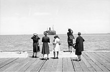 Five women standing at the edge of a dock. A large ship is sailing away from them.