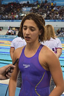 Pignatiello trägt einen lila Badeanzug. Sie spricht in ein Mikrofon, hinter ihr ist ein Schwimmbecken.