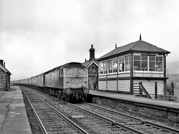 A British Rail Class 47 locomotive no.47561 hauling diverted inter-city West Coast train passes Garsdale signal box and disused station on the Down Ma