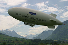 An airship flies from right to left, nose up, against a background of mountains