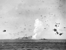 12 août 1942 : Le HMS Indomitable en feu après son bombardement. On aperçoit le croiseur antiaérien HMS Charybdis en premier plan