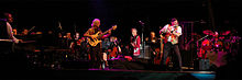 A group of musicians playing various instruments on a stage, including a flute, guitars, piano and drum set. Additional instruments, orchestra members and audio equipment can be seen on stage and in the background.