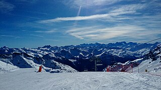 Photographie de pistes de ski, damées ; en face on voit les montagnes bleues enneigées.