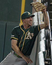 Een man in een donkergroen baseballshirt en pet met het Oakland Athletics A-logo op de voorkant probeert een honkbal te vangen.