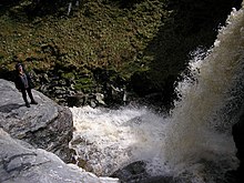 Schaumiges Wasser fällt aus einer Höhe von etwa 10 m in ein turbulentes Wasserbecken, das von Felsen umrahmt ist