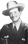 Мъж, облечен в бяла каубойска шапка и тъмно яке, широко усмихнат и държащ китара