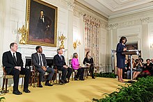 Five people seated on a stage under a large portrait of Lincoln while Michelle Obama stands on the stage speaking.