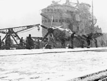 The flight deck of an aircraft carrier during a snowstorm, with the ship's superstructure in the background. A propeller aircraft is sitting on the snow-covered flight deck, and is being worked on by several people.