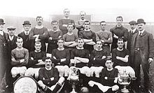 A black and white photograph of a football team in alternating light and dark shirts with three trophies at the front.