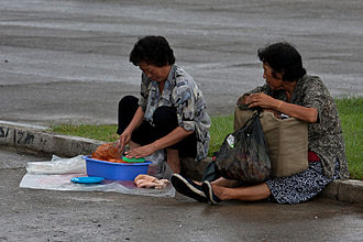 Two elderly North Korean women sitting on street. One of them is preparing food.