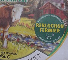 Gros plan sur la pastille verte et jaune figurant sur l'emballage d'un reblochon fermier, à l'arrière plan figue sur l'emballage le dessin d'un chalet et d'une vache de race montbéliarde.